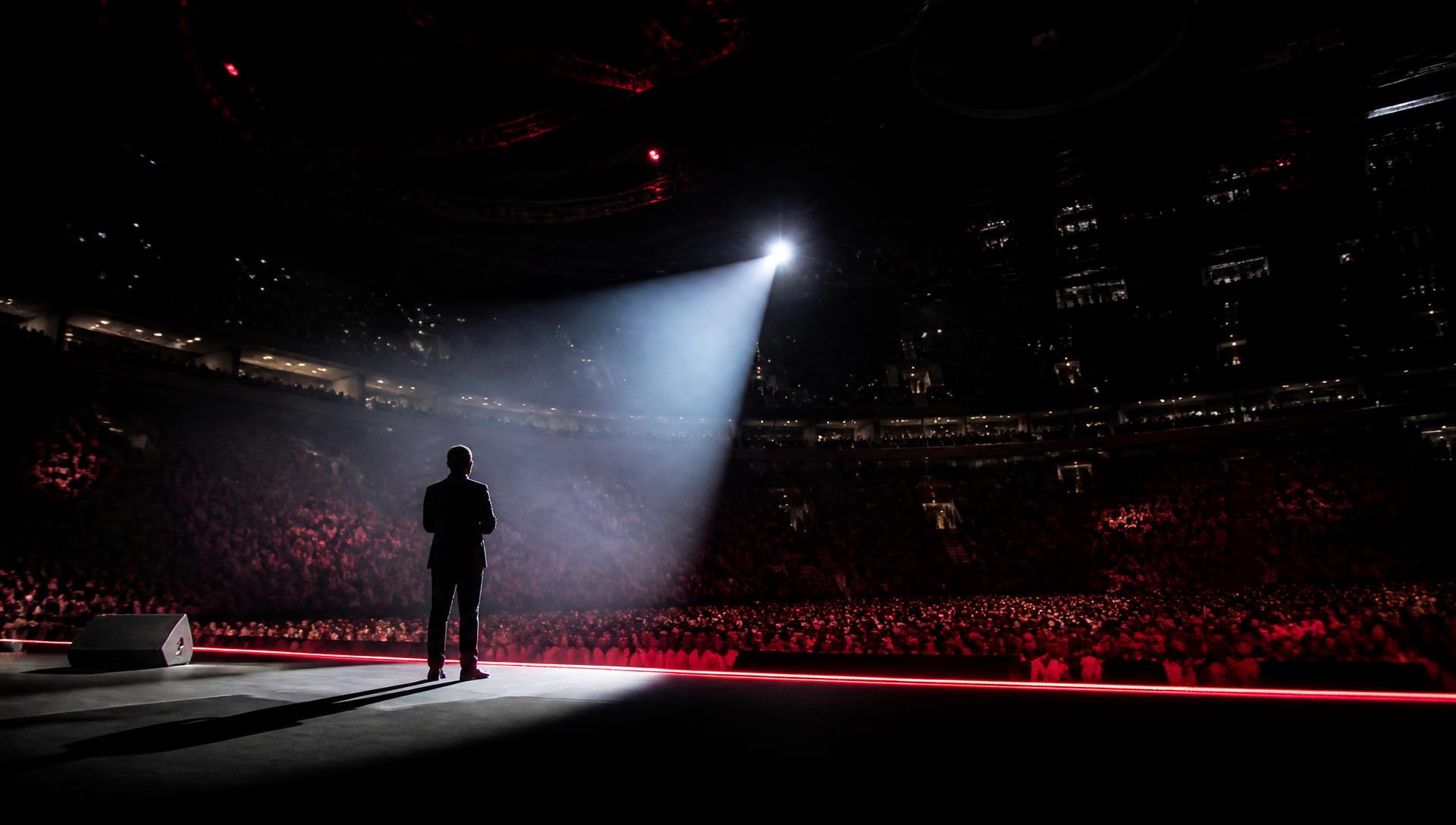 A speaker alone in a spotlight on a massive arena stage in front of thousands